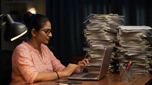 Focused woman working on laptop with huge stack of paperwork, late at night. Overworked adult with technology and workload at desk in office.