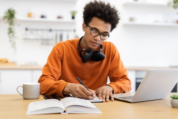 A young Black man wearing glasses writes in a notebook while studying at home with a laptop and coffee.