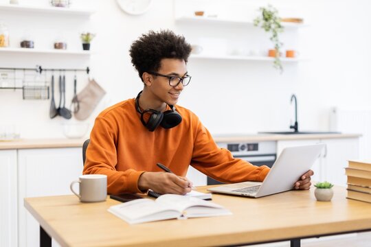 A young man with glasses studies at a table with a laptop, notebook, and coffee mug in a bright kitchen setting.