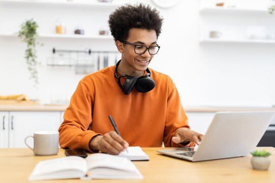 A young man with glasses works on a laptop while taking notes in a bright kitchen setting.