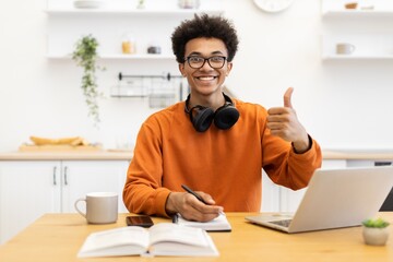 A cheerful young man with glasses smiles and gives a thumbs up while working on his laptop at home.