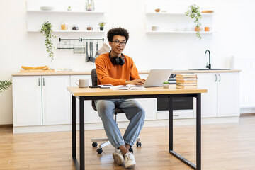 A young man with glasses sits at a desk, working on a laptop in a bright, modern kitchen setting.
