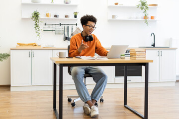 A young man with glasses is working on his laptop at a table in a modern kitchen setting.