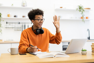 A young man with glasses waves enthusiastically during a video call on his laptop, likely in a home office setting.