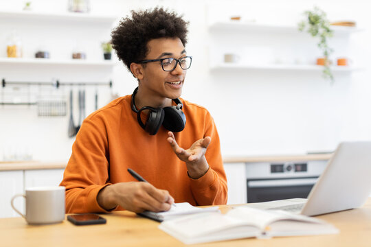 A young man with glasses is engaged in a video call, taking notes on a notepad while working from home. - Powered by Adobe