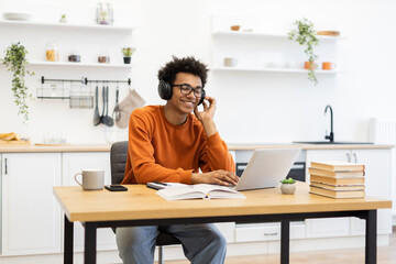 A young man with headphones smiles while working on a laptop at a wooden table in a bright kitchen setting.