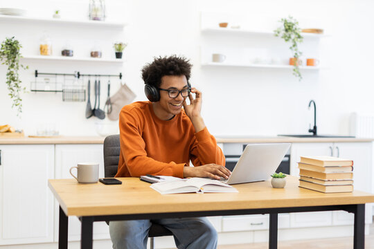 A young man with glasses and headphones smiles while working on a laptop at a wooden table in a modern kitchen setting.