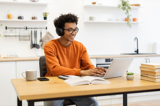 A young man with glasses and headphones smiles while working on his laptop at a wooden table in a bright kitchen setting.