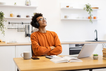 A young man wearing headphones smiles while listening to music at his kitchen table, with a laptop and coffee nearby.