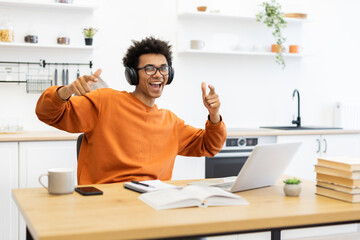 A joyful man with headphones points at the camera while working on a laptop at home.