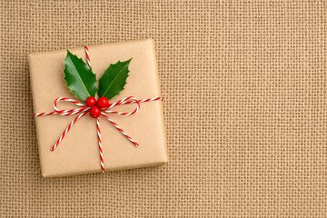 Christmas gift wrapped in kraft paper with holly and red berries on burlap background, eco friendly holiday packaging concept