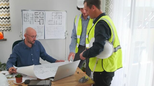 caucasian site supervisor leaning over desk to clarify blueprint details with seated client, safety helmet on table, laptops open, measuring tape and notes visible, cooperative problem solving mood