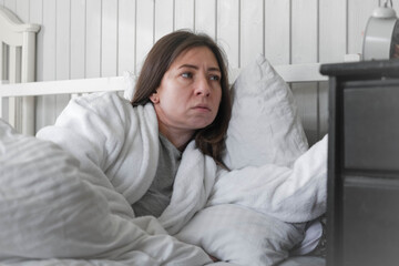 A woman, wrapped in a soft robe, with a wary expression on her face, looking at the alarm clock, among light pillows and a calm interior, anticipating the silence of the early morning. Copy space