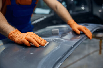 Auto technician sanding car body panel with orange gloves in workshop