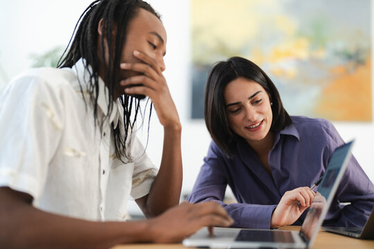 Two diverse colleagues collaborate on a laptop in a bright office. The woman smiles while pointing at the screen, and the man types, engaged in their work.