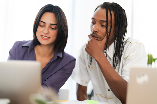 Two diverse young professionals collaborate on a project, looking at a laptop screen. They are engaged in discussion and problem-solving in a modern office setting.