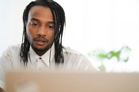 A young Black man with braided hair and a beard intently looks down at a laptop. He is engaged in work or study in a bright, modern setting.