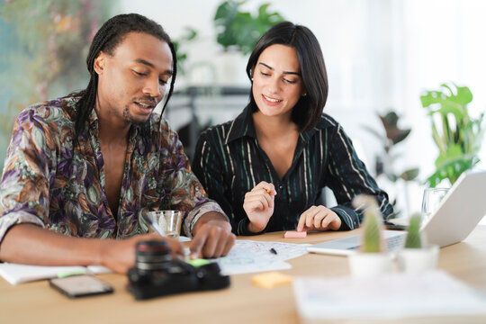 Two diverse professionals collaborate at a desk, reviewing documents. The man points at papers while the woman smiles, holding a pencil. They share ideas and work together.
