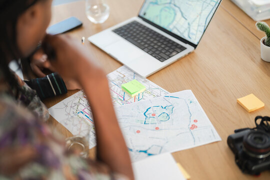 An individual plans a route or project at a desk, surrounded by paper maps, a laptop displaying a map, and sticky notes. A camera rests nearby.