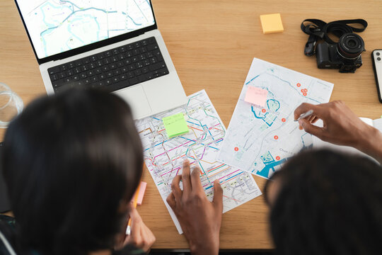 Two people collaborate on travel plans, examining physical maps and a laptop displaying a digital map. They are organizing their itinerary with sticky notes, preparing for an adventure.