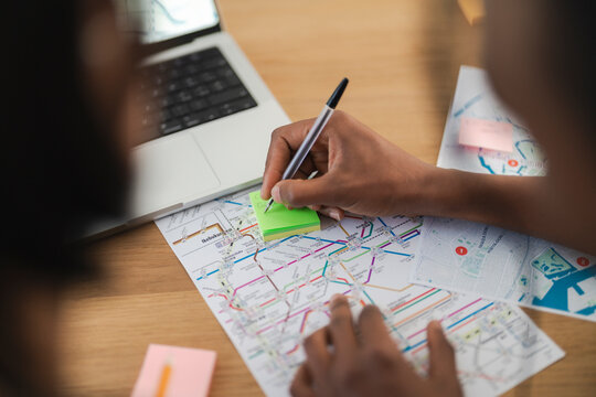 A person writes notes on a green sticky pad over a detailed transport map, with a laptop open on the table. This depicts careful planning or route analysis.