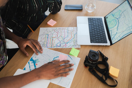 Two individuals are at a wooden table, studying maps and a laptop with a digital map. They are planning a journey, with sticky notes and a camera nearby for their adventure.