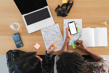 Two people meticulously plan their route, comparing a smartphone's digital map with a large printed map on a wooden table. They are taking notes and using a laptop.