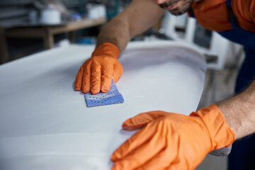 Craftsman sanding surfboard in workshop with hands with orange gloves finishing board