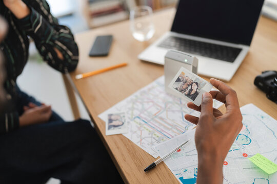 A person holds an instant photo of two friends, reminiscing about travel memories. Maps, a laptop, and a printer are on the table, suggesting trip planning.