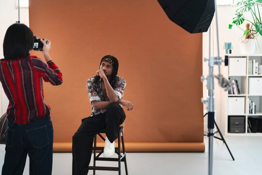 A female photographer captures a portrait of a male model posing on a stool in a professional studio. The model wears a patterned shirt and bandana. Studio lights are set up.
