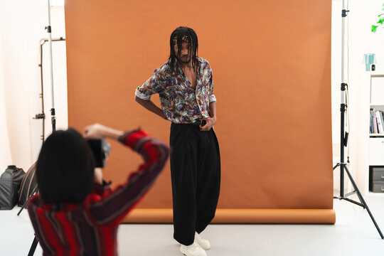 A photographer captures a male model posing in a studio. The model, with dreadlocks and a patterned shirt, stands against a brown backdrop during a photoshoot.