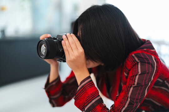 A young woman with dark hair holds a modern digital camera to her eye, focusing intently on a subject. She is taking a photograph indoors, capturing a special moment.
