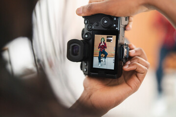 A photographer holds a digital camera, reviewing a shot of a female model posing on a stool. The camera screen shows the model with focus and exposure details.