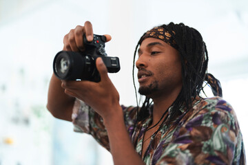 A young Black man with dreadlocks and a bandana holds a camera, focusing intently on his subject. He presses a button, capturing a moment or adjusting camera settings.
