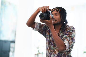 A young man with dreadlocks and a patterned shirt holds a digital camera, looking through the viewfinder. He is focused on capturing an image, showcasing his photography skills.