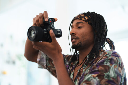 A young Black man with dreadlocks and a bandana holds a camera, focusing intently on his subject. He presses a button, capturing a moment or adjusting camera settings.