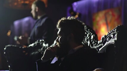 Mysterious man with a drink sitting in a dark cinematic room. Dramatic mafia-style scene with men in low light. Bearded man holding a glass in a shadowy interior. Dark cinematic mood with rich man.