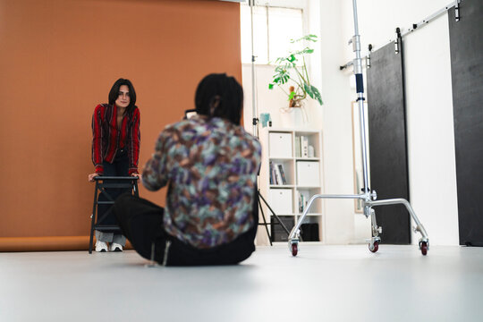 A female model poses for a photographer in a bright studio. The photographer sits on the floor, capturing the model leaning on a stool against a brown backdrop.