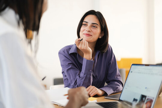 Two young women collaborate in a bright office. One woman smiles, listening attentively with a pen. The other is writing in a notebook during their productive meeting.