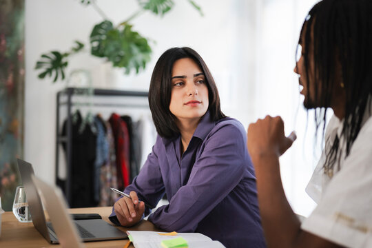Two diverse professionals are engaged in a focused discussion at their modern office workspace. The woman listens attentively, holding a pen, as her male colleague shares ideas.