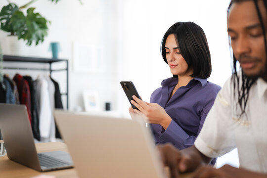 A focused woman uses her smartphone at a desk with a laptop, while a man works on his computer in the foreground of a modern office setting.