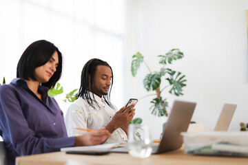 Two diverse professionals collaborate in a bright office. A woman writes in a notebook, while a man engages with his mobile phone. Laptops and plants are visible on the desk.