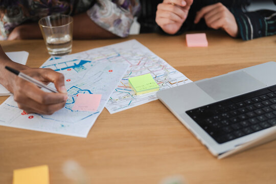 Two individuals collaborate, planning a route or project using a detailed map. One person marks locations with a pen, while sticky notes indicate key points. A laptop sits nearby.