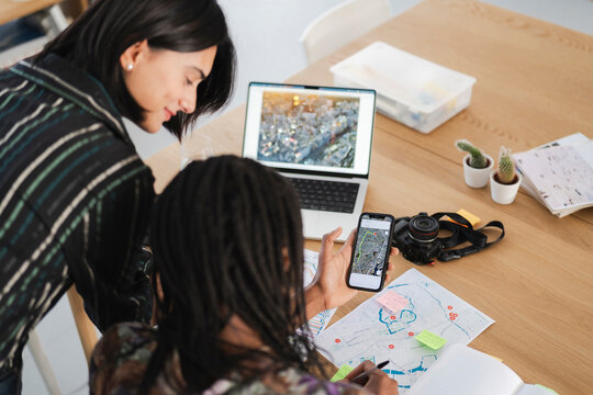 Two women collaborate on travel plans, using a smartphone map, laptop, and physical map. They are organizing a trip, possibly to Tokyo, combining digital and traditional tools.