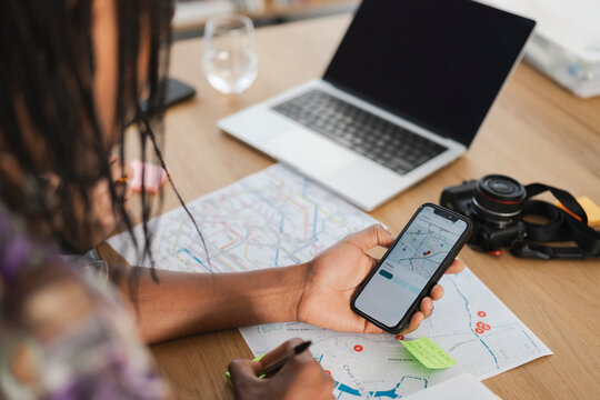 A person plans a trip, consulting a digital map on a smartphone and a physical map. They write notes, organizing their travel itinerary with a laptop and camera nearby.