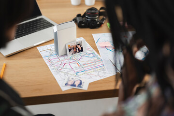 Two individuals review maps and instant photos on a wooden desk. A portable photo printer and laptop are nearby, capturing and sharing travel memories and plans.
