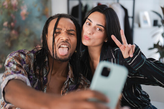 A diverse young couple takes a playful selfie. The man makes a funny face with his tongue out, while the woman pouts and shows a peace sign. They are enjoying a fun moment together. - Powered by Adobe
