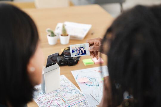Two people share a printed instant photo, looking at it together. Maps and a camera are on the table, suggesting travel planning. They enjoy the moment and memories.