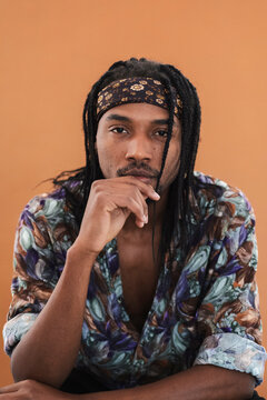 A young Black man with dreadlocks and a patterned headband looks directly at the camera. He wears a colorful shirt and has a thoughtful expression, with his hand near his chin.