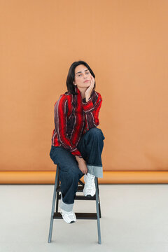 Young woman with dark hair sits on a step stool in a studio. She wears a striped shirt, jeans, and white sneakers, posing with her hand on her chin.